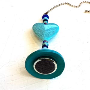 May include: A decorative ceiling fan pull chain with a turquoise heart-shaped charm. The charm is connected to a teal disc with a silver accent and a dark brown center. The chain is silver.