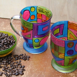 Pode incluir: Duas canecas de caf&eacute; de vidro coloridas com desenhos geom&eacute;tricos em tons de azul, verde, rosa, laranja e roxo. As canecas t&ecirc;m al&ccedil;as transparentes e est&atilde;o sobre uma superf&iacute;cie de madeira.