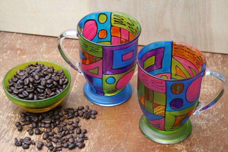 Pode incluir: Duas canecas de caf&eacute; de vidro coloridas com desenhos geom&eacute;tricos em tons de azul, verde, rosa, laranja e roxo. As canecas t&ecirc;m al&ccedil;as transparentes e est&atilde;o sobre uma superf&iacute;cie de madeira.