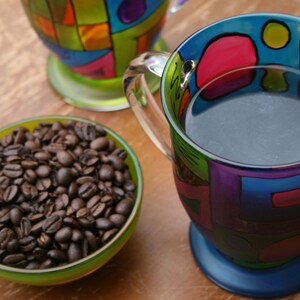 Pode incluir: Uma caneca colorida com um design azul, verde, roxo e vermelho. A caneca est&aacute; cheia de caf&eacute; escuro e tem uma al&ccedil;a. Uma tigela de gr&atilde;os de caf&eacute; est&aacute; no fundo.