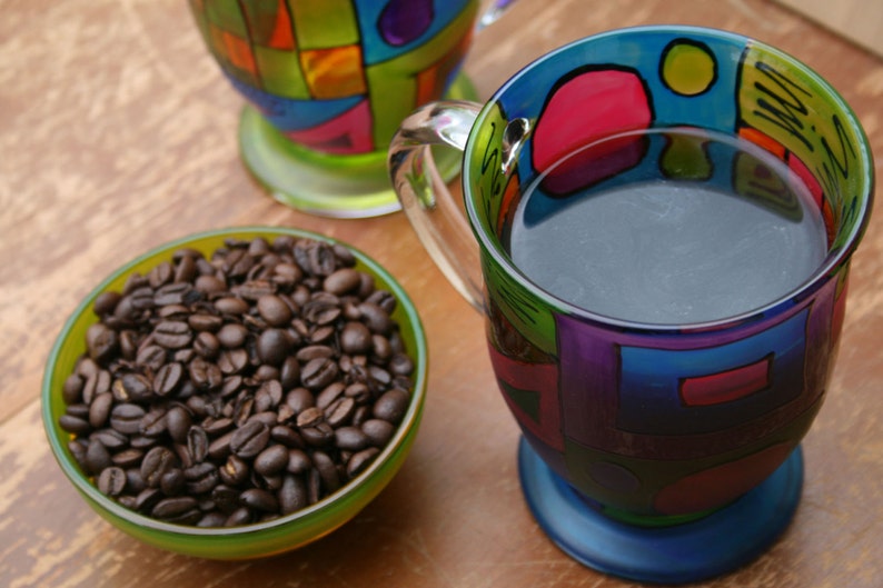 Pode incluir: Uma caneca colorida com um design azul, verde, roxo e vermelho. A caneca est&aacute; cheia de caf&eacute; escuro e tem uma al&ccedil;a. Uma tigela de gr&atilde;os de caf&eacute; est&aacute; no fundo.