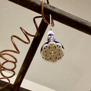 May include: A decorative ornament with a white, snowflake-like design over a dark blue base. The ornament is suspended from a silver hook and features a beaded top. The background includes copper tubing and a wooden structure.