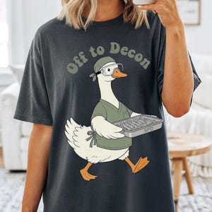 Könnte beinhalten: Ein anthrazitfarbenes Kurzarm-T-Shirt mit einer Grafik einer weißen Gans, die ein grünes OP-Oberteil, eine passende Haube und eine Schutzbrille trägt. Die Gans hält ein Metalltablett, darüber steht der Text Off to Decon gedruckt.