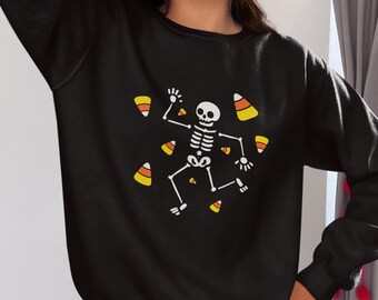 May include: Black sweatshirt featuring a white skeleton graphic with candy corn accents. The skeleton is in a playful pose, surrounded by scattered candy corn. A fun and festive Halloween-themed design.
