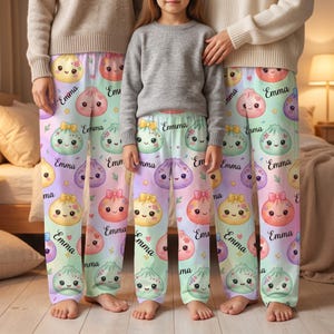 May include: Matching pajama pants featuring a repeating pattern of colorful, smiling cartoon dumplings with bows and the name Emma printed in black script. The background is a soft pastel gradient of light purple, blue, and green with small stars.