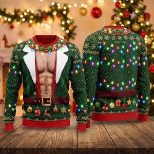 Puede incluir: Un suéter verde de manga larga con un gráfico impreso de un torso desnudo con borde de piel blanca, un cinturón marrón con hebilla dorada y luces navideñas de colores. El diseño incluye adornos colgantes, un reno y copos de nieve.