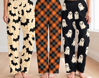 May include: Three pairs of lounge pants with elastic waistbands. The left pair is cream with black bat silhouettes. The center pair features an orange, black, and gray diagonal plaid pattern. The right pair is black with white ghost illustrations.