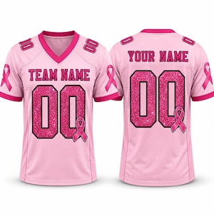 May include: A light pink sports jersey with a V-neck, short sleeves, and mesh side panels. The front features the text TEAM NAME and the number 00 in glittery pink, with a pink ribbon graphic. The back displays YOUR NAME and the number 00.
