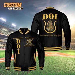 Könnte beinhalten: Schwarze Bomberjacke mit goldenen Zierleisten und dem Text "DOI" auf der Vorder- und Rückseite. Auf der Rückseite der Jacke befindet sich eine Illustration und die Worte "Inner Women Mind Body Spirit". Auf der Vorderseite der Jacke steht der Text "Dt LaQuenette Bromell Kaaba Court #127 Oasis of Conway Desert of South Carolina".