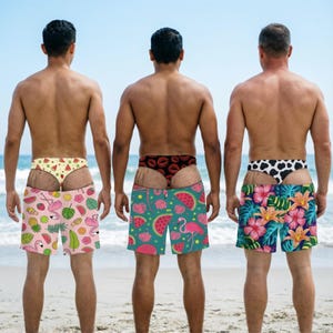 Op de afbeelding: Drie zwembroeken met verschillende patronen. De eerste is roze met flamingo's en fruit. De tweede is blauwgroen met watermeloenen en flamingo's. De derde heeft een tropische bloemenprint met hibiscus en palmbladeren.