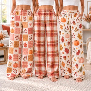 May include: Three pairs of wide-leg lounge pants featuring autumn-themed patterns. The first pair has a patchwork design with pumpkins, leaves, and hearts. The second pair has a red and white plaid pattern. The third pair features pumpkins, acorns, and leaves.