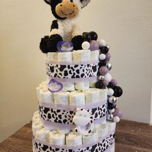 May include: A three-tiered diaper cake decorated with a plush cow, pacifier, and cow-print ribbon. The cake is adorned with decorative beads in black, white, silver, and purple. A cow-shaped rattle is attached to the side.