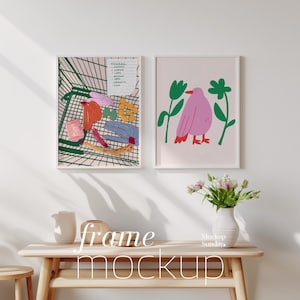 Puede incluir: Dos ilustraciones enmarcadas en una pared blanca. El marco de la izquierda muestra un carro de la compra lleno de comestibles coloridos. El marco de la derecha muestra un pájaro rosa con hojas verdes. Los marcos están sobre una mesa de madera clara con un jarrón de flores blancas.