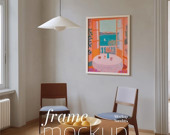Op de afbeelding: Een ingelijste kunstdruk met een kleurrijke kustscène hangt aan een grijze muur. Twee houten stoelen met blauwe zittingen en een houten tafel staan voor de kunst. Een witte hanglamp hangt aan het plafond. De tekst "frame mockup" staat op de tafel.