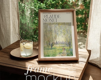 May include: A framed print of a Claude Monet painting, with the artist's name and the words "Mockup Sunday" visible. The frame is brown and sits on a wooden table. A glass of lemonade and a small plate are also on the table.