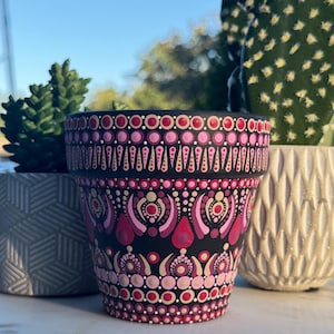 May include: A black ceramic flower pot with a hand-painted design in shades of pink, red, and beige. The pot features a repeating pattern of dots and teardrop shapes. The pot is empty and ready for planting.