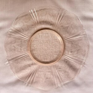 Puede incluir: Un plato de vidrio transparente y rosado con un patrón floral y un borde festoneado. El plato tiene un centro elevado y líneas radiales que se extienden desde el centro hasta el borde. El plato tiene aproximadamente 25 cm de diámetro.