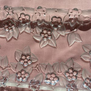May include: Clear, floral-shaped embellishments with silver-colored centers and leaf accents. The decorative trim is arranged in rows, creating a delicate, transparent design. The background is a soft pink fabric.