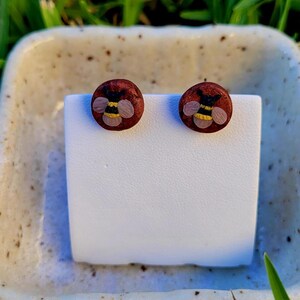 May include: A pair of round stud earrings with a dark red background. Each earring features a detailed bee design with yellow and black stripes. The earrings are displayed on a white stand, set against a blurred green background.