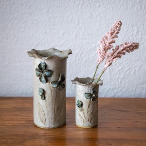 May include: Two ceramic vases with a floral design. The taller vase has a ruffled top and is decorated with green flowers and leaves. The smaller vase also has a ruffled top and holds two stems of pink flowers. Both vases are on a wooden surface.