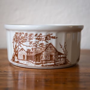 May include: A white ceramic bowl featuring a brown illustration of a rural cabin. The bowl has a wide, round shape with a slightly flared rim. The cabin scene includes a house, trees, and a fence.
