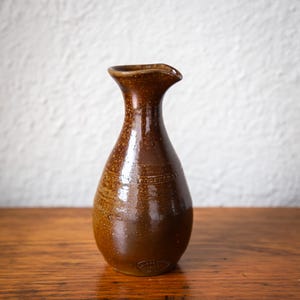 May include: A brown ceramic vase with a narrow neck and a flared spout. The vase has a rounded body and a textured surface with colour variations. A decorative band encircles the upper body. The vase is likely handmade.