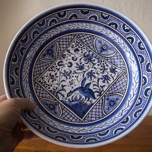 May include: A round ceramic plate with a blue and white floral design. The plate features a central diamond-shaped pattern with a bird and floral motif. The rim has a repeating pattern of blue and white shapes. The plate is held by a hand.