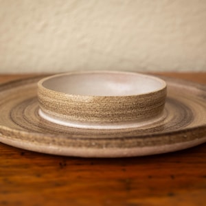 May include: A ceramic serving set, comprising a shallow bowl set within a large, round platter. The set has a natural, earthy appearance with brown and tan hues. Suitable for serving appetizers or small dishes. Measurements in cm.