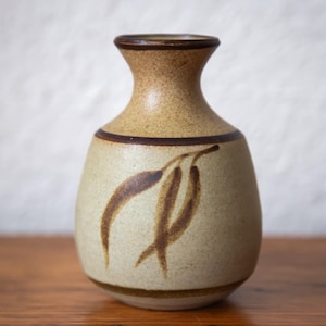 May include: A ceramic vase with a speckled tan body and a narrow neck. The vase has a brown band around the neck and base, with a brown leaf design on the front. The vase is displayed on a wooden surface.