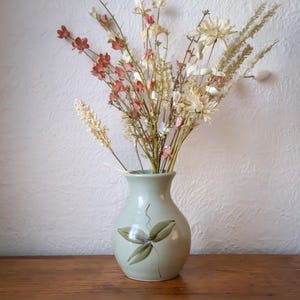 May include: A pale green ceramic vase with a painted floral motif contains a collection of dried flowers. The arrangement features white, pink, and beige flowers and stems. The vase rests on a wooden surface against a textured white wall.
