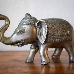 May include: A detailed silver-toned elephant figurine with embossed patterns on its back and head. The elephant has a curved trunk and large ears. The figurine is displayed on a wooden surface.