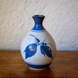 May include: A small, white ceramic vase with a blue floral design. The vase has a rounded body, a narrow neck, and a dark blue rim. The blue design features leaves and berries.