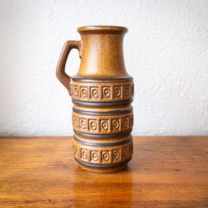 May include: A brown ceramic jug with a handle. The jug has a textured surface and is decorated with geometric patterns. The jug is approximately 25 cm tall and sits on a wooden surface.