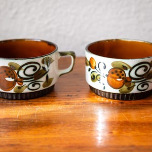 May include: Two ceramic mugs with a white base, brown interior, and a decorative floral pattern in orange, brown, and green. The mugs have small handles and sit on a wooden surface.