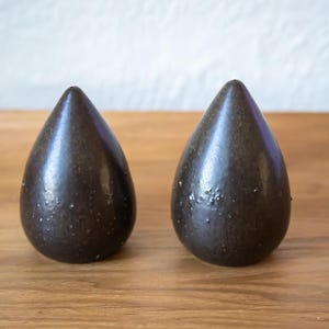 May include: Two dark brown, teardrop-shaped objects with a textured surface. The objects are placed side-by-side on a wooden surface. The background is a light-coloured wall.