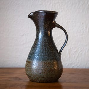 May include: A ceramic jug with a dark brown top, fading into a blue-green and brown speckled body. It features a curved handle and a pouring spout. The jug is displayed on a wooden surface.