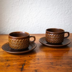 May include: Two brown ceramic teacups with matching saucers. The cups and saucers have a repeating arched pattern. The cups have a handle on the side. The set is on a wooden surface.