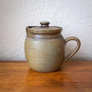 May include: A handmade ceramic jar with a lid and handle. The jar is a muted olive green with a darker brown rim and lid. The handle is curved, and the jar sits on a wooden surface.