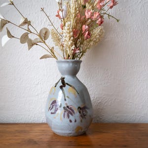 May include: A pale blue ceramic vase with a narrow neck and a rounded body, adorned with purple and yellow floral patterns. The vase contains a display of dried flowers and foliage. The vase sits on a wooden surface.