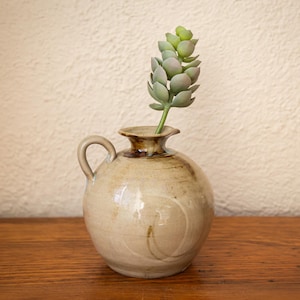 May include: A small, round, beige ceramic pitcher with a single side handle and a flared rim. The surface features subtle brown glaze streaks and swirling patterns. A single artificial succulent stem with pale green, rounded leaves sits inside.
