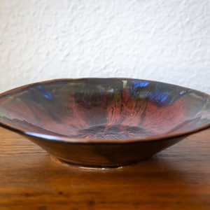 May include: A handmade ceramic bowl with a wide, shallow design. The bowl has a dark brown exterior and a vibrant interior with shades of red, black, and blue. The bowl is placed on a wooden surface.