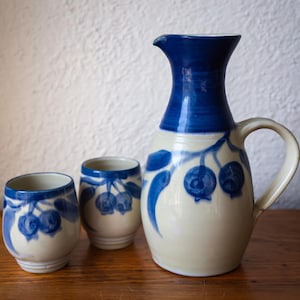 May include: A ceramic jug and two matching cups. The jug has a dark blue top and handle, with a cream-coloured body decorated with blue berry designs. The cups have a similar design, with blue rims and berry motifs. The set is on a wooden surface.