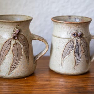 May include: Two handmade ceramic mugs with handles. The mugs are beige with brown leaf and bud designs. The mugs are sitting on a wooden surface. The mugs are likely used for drinking coffee or tea.