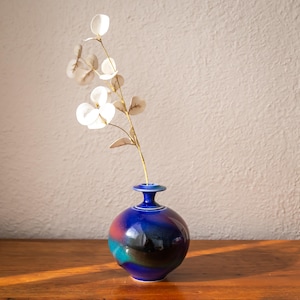 May include: A small, round, blue ceramic vase with a narrow neck and a white rim. The vase has a glossy finish with bands of red, teal, and black. A sprig of white flowers with beige leaves is placed inside the vase.