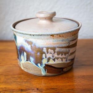 May include: A handmade ceramic jar with a lid. The jar is cylindrical with a light beige base and lid. The exterior features a blend of brown, blue, and white glaze, creating a unique, artistic appearance. The jar is approximately 13 cm in height.
