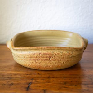 May include: A square ceramic baking dish with a light brown glaze. The dish has rounded corners, a textured surface, and small handles. Decorative etched lines are visible. It sits on a wooden surface. Suitable for baking.