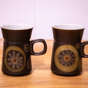 May include: Two ceramic mugs. The dark brown exterior contrasts with the white interior. Each mug has a floral design in yellow, brown, and grey. The mugs have handles and are on a wooden surface.