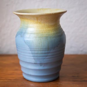 May include: A handcrafted ceramic vase with a flared rim. The vase displays a gradient of colours, shifting from yellow at the top to blue at the base. The vase has a textured, ridged design and is on a wooden surface.