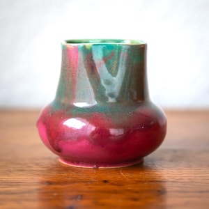 May include: A small ceramic vase with a rounded base and a slightly flared top. The vase has a deep magenta base that transitions to shades of green and teal near the rim. The glossy finish enhances the colours. A decorative piece for the home.
