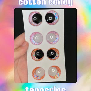 Op de afbeelding: Een set van acht poppenogen in de kleuren suikerspin en mandarijn. De ogen hebben zwarte pupillen en glitteraccenten. De woorden "cotton candy" en "tangerine" staan boven en onder de ogen.